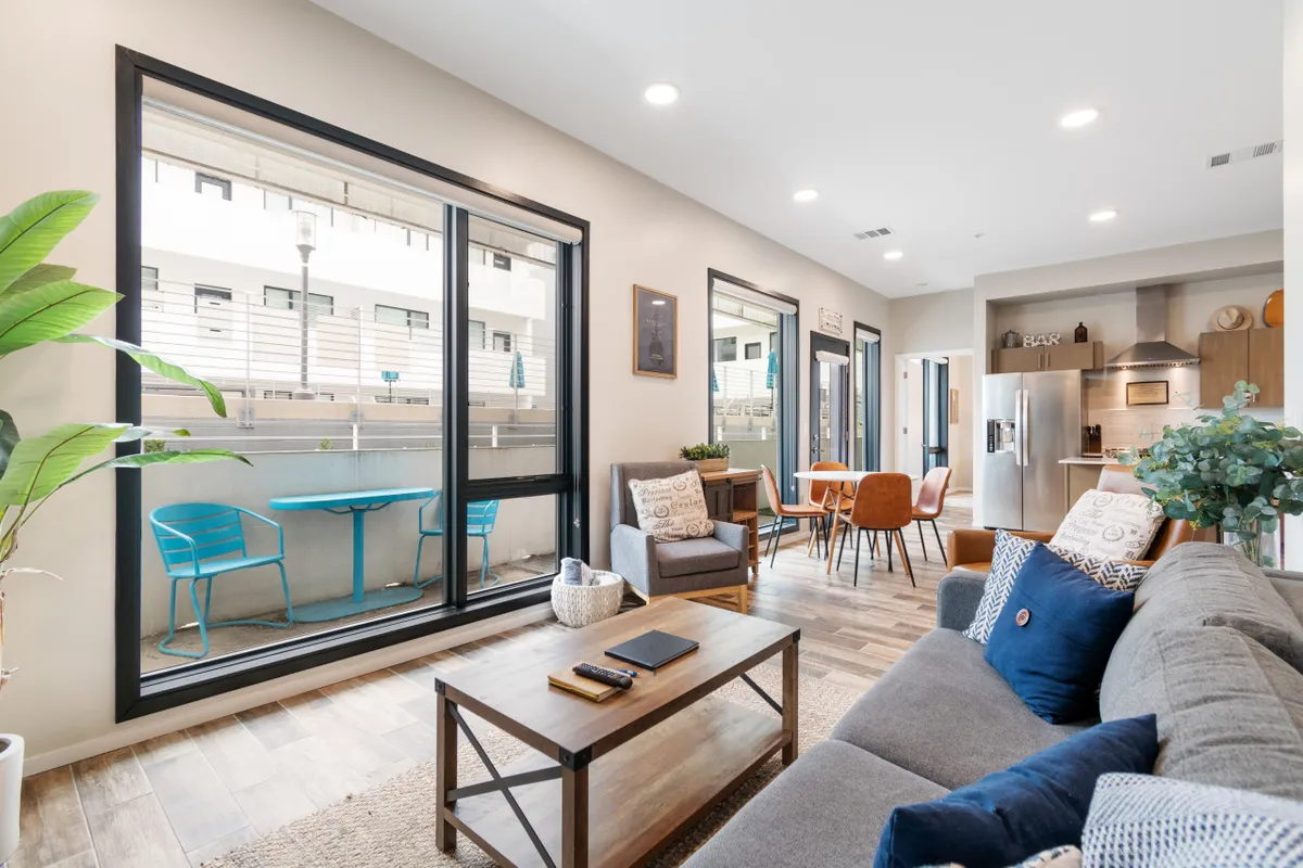 A spacious open-plan living, dining, and kitchen area bathed in sunlight from large windows. A comfortable sofa anchors the living space, with a stylish dining table adjacent and a modern kitchen with island seating in the background. The overall mood is airy and welcoming, with plenty of room for gatherings and relaxation.