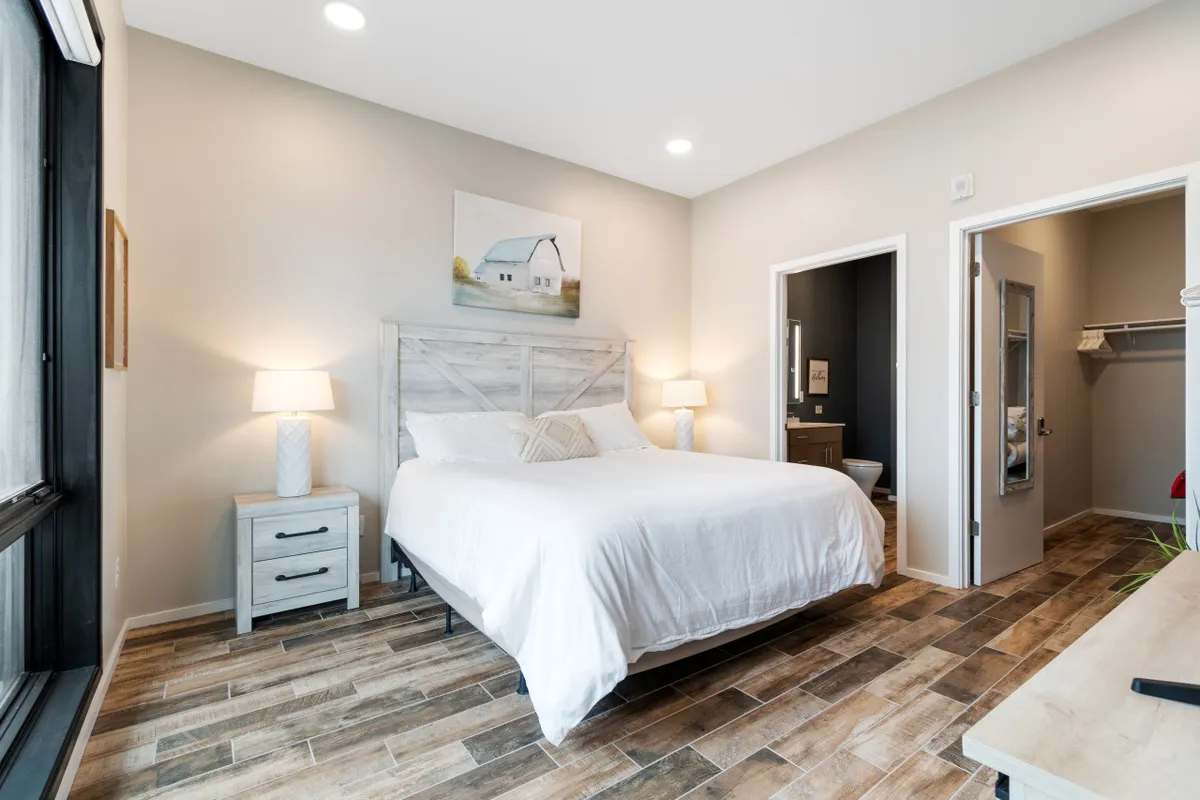 A serene bedroom featuring a queen bed adorned with plush bedding, a nightstand with a lamp for evening reading, a closet door that hints at generous storage, and soft natural light enhancing the peaceful atmosphere. Painted artwork above the bed adds a touch of personal style.