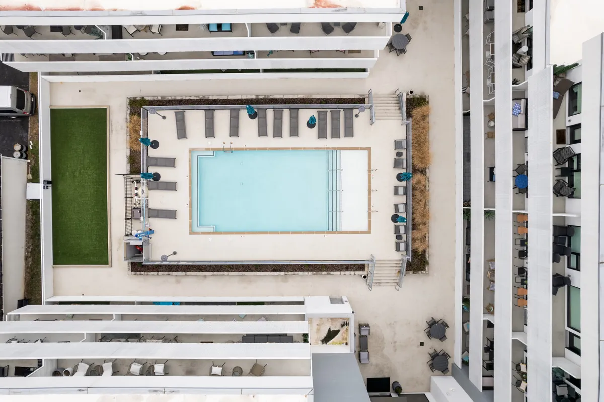 A vibrant exterior scene showcasing a sparkling swimming pool surrounded by comfortable lounge chairs and an outdoor bar area. The relaxed atmosphere is enhanced by the presence of people sunbathing and socializing, with the pool reflecting soft sunset light.