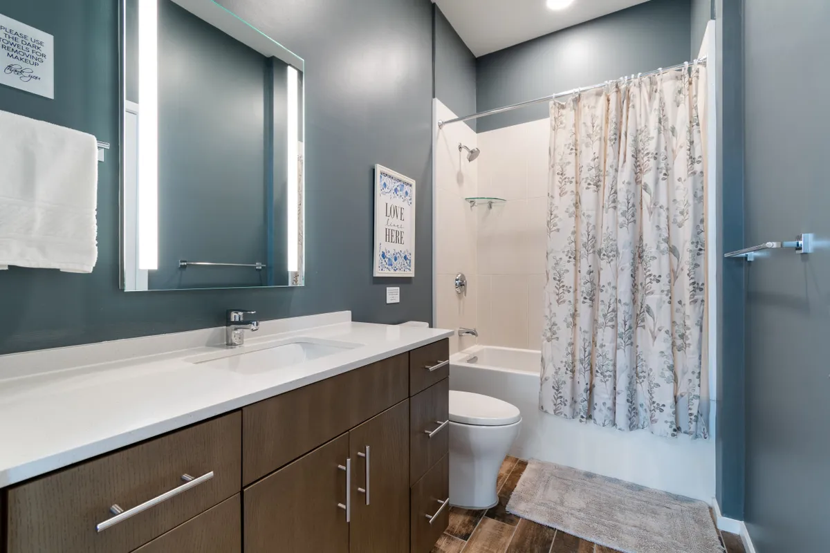 A bright bathroom with a vanity, large mirror, shower area, and decorative wall art. Morning sunlight filters through a window, creating a serene spot for daily routines or a rejuvenating bath.