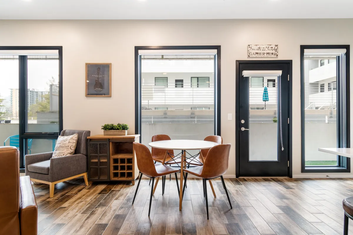 A cozy dining table positioned by sun-filled windows, complemented by an armchair and decorative plant. The setup suggests a comfortable environment for group meals, casual gatherings, or catching up over coffee, with plenty of natural light.