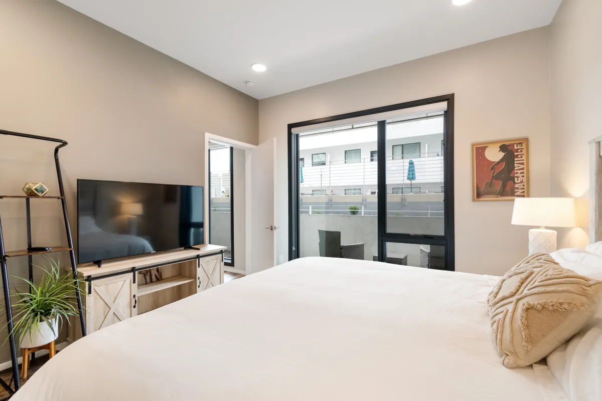 A serene bedroom scene with a bed facing a smart TV, large window, and lamp. Natural light spills through the window, creating an inviting spot for relaxing after work, reading, or enjoying a movie. The setup hints at spaces suitable for both relaxation and informal work-from-bed moments.
