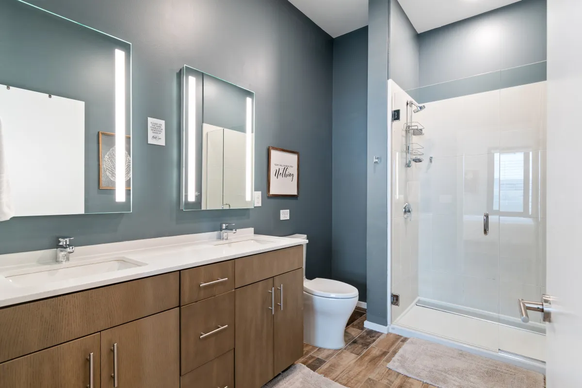 A modern bathroom showcasing dual LED-lit mirrors above a spacious double vanity, a glass-enclosed walk-in shower, and calm, inviting finishes. The layout supports both privacy and comfort for morning routines or relaxation.