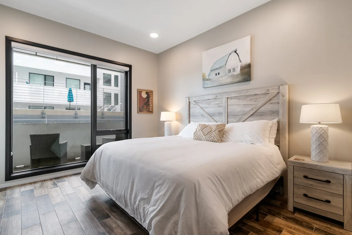 A bright, serene bedroom featuring a comfortable bed positioned beside a large window, with art on the wall and stylish nightstands topped with lamps. The space is filled with natural light and offers a peaceful spot for relaxation or enjoying a morning coffee.