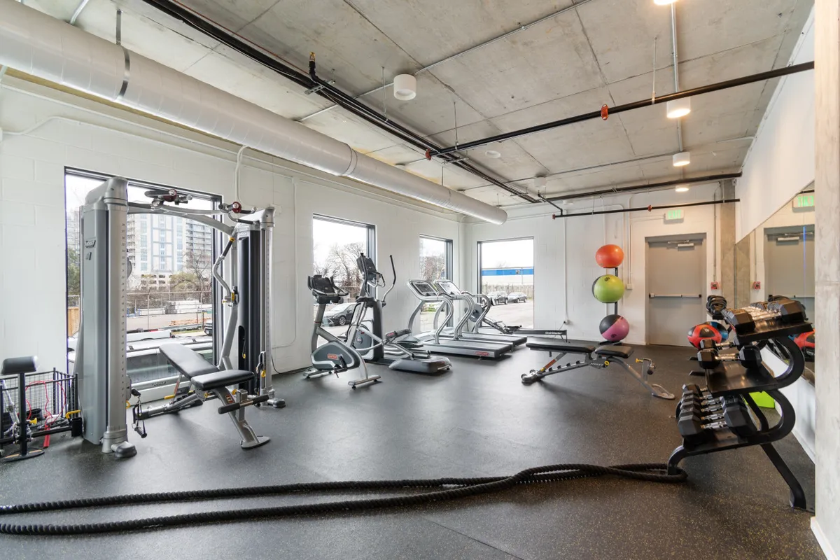 A well-equipped fitness center featuring exercise machines, free weights, and exercise balls, all bathed in natural light from large windows. The modern setup is designed to motivate residents to maintain their fitness routines.